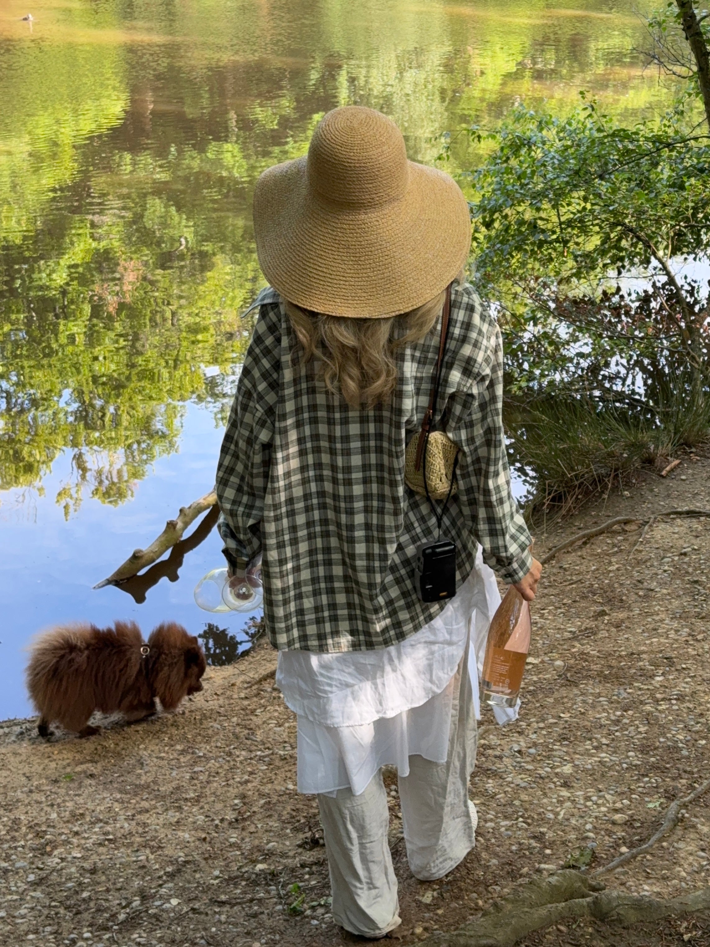 Model walking a dog by a lake, wearing a checkered shirt and wide-brimmed hat holding an unopened wine bottle