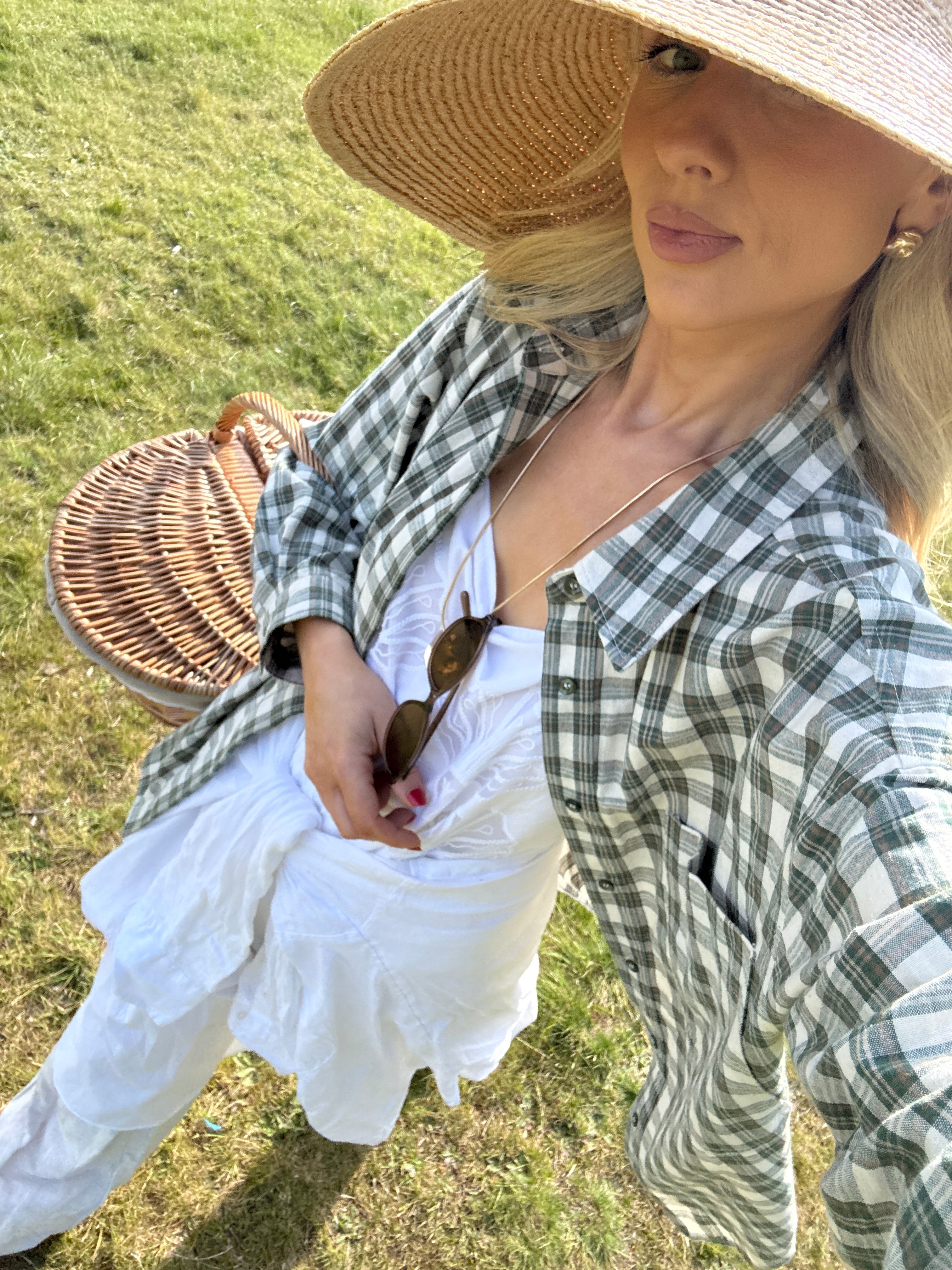 Woman in a checkered shirt and white dress holding a picnic basket in a grassy field