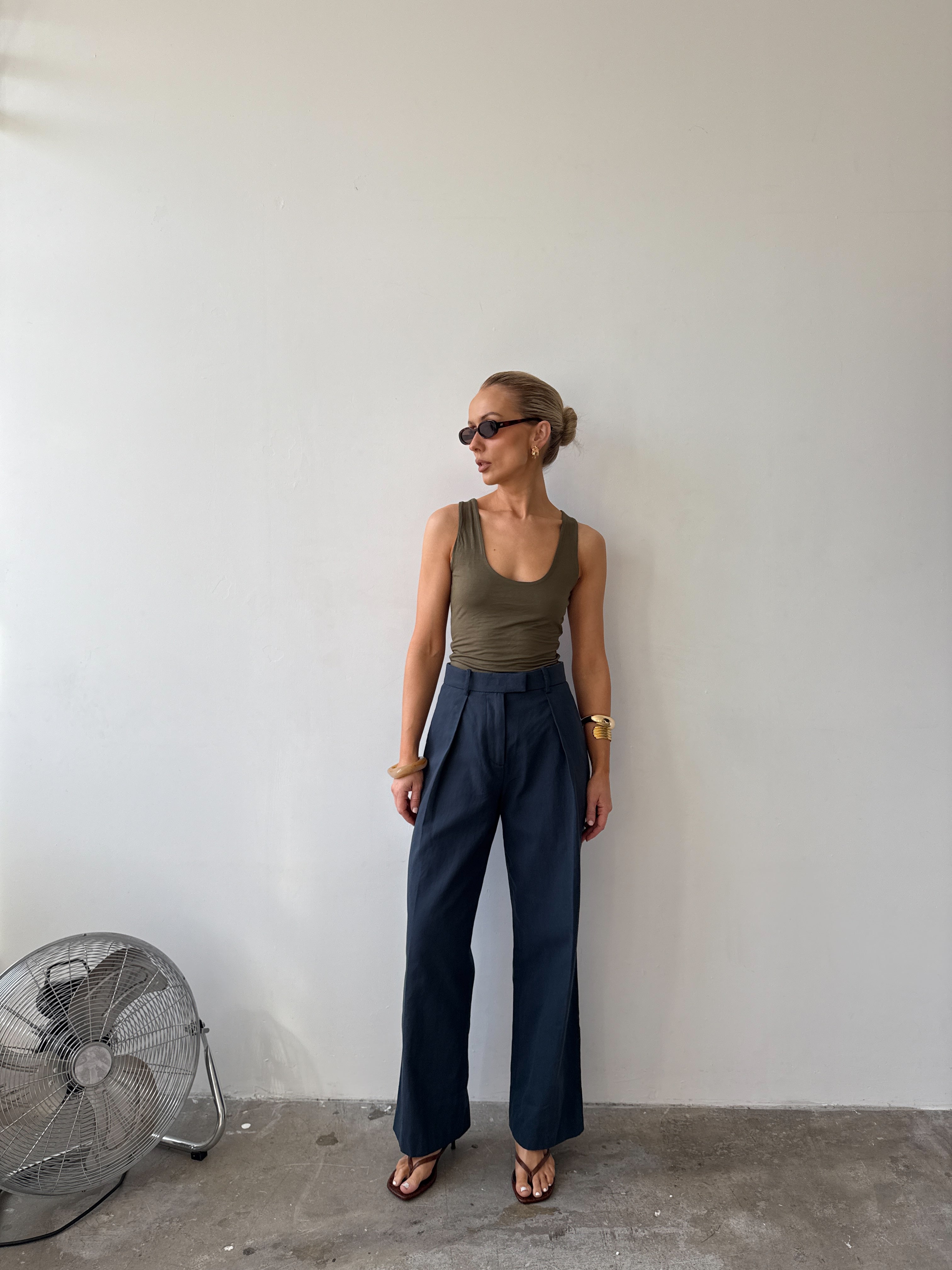 Model wearing ink blue tailored trousers with front pleats styled with a green tank top, with sunglasses on and flat sandals.