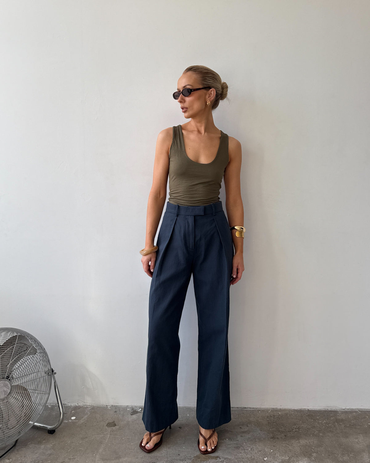 Model wearing ink blue tailored trousers with front pleats styled with a green tank top, with sunglasses on and flat sandals.