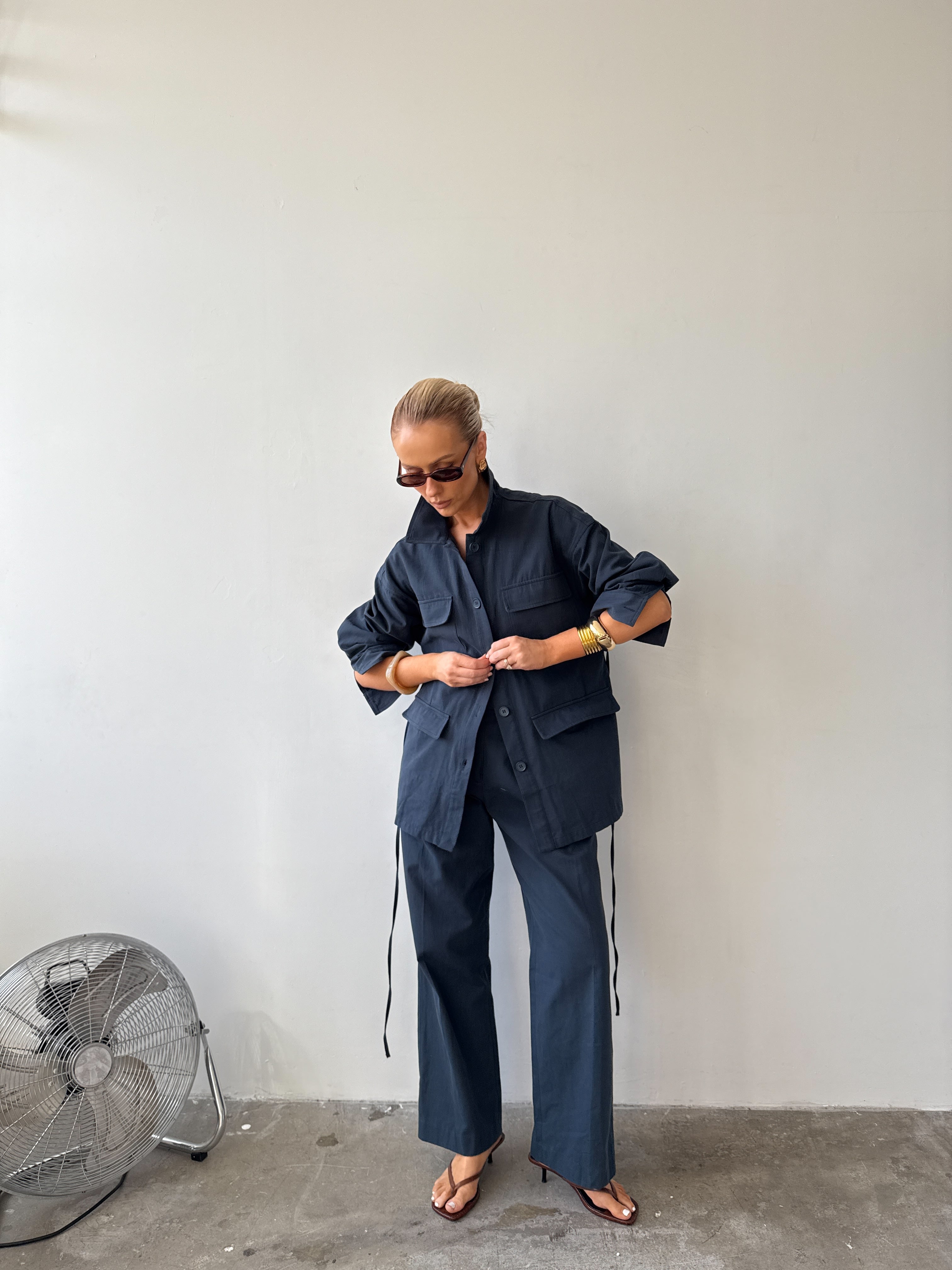 Model wearing navy two‑piece set (The Ink Blue Field Jacket and Day Trousers) while standing against a plain pale wall, wearing sunglasses, gold bracelets and sandals.