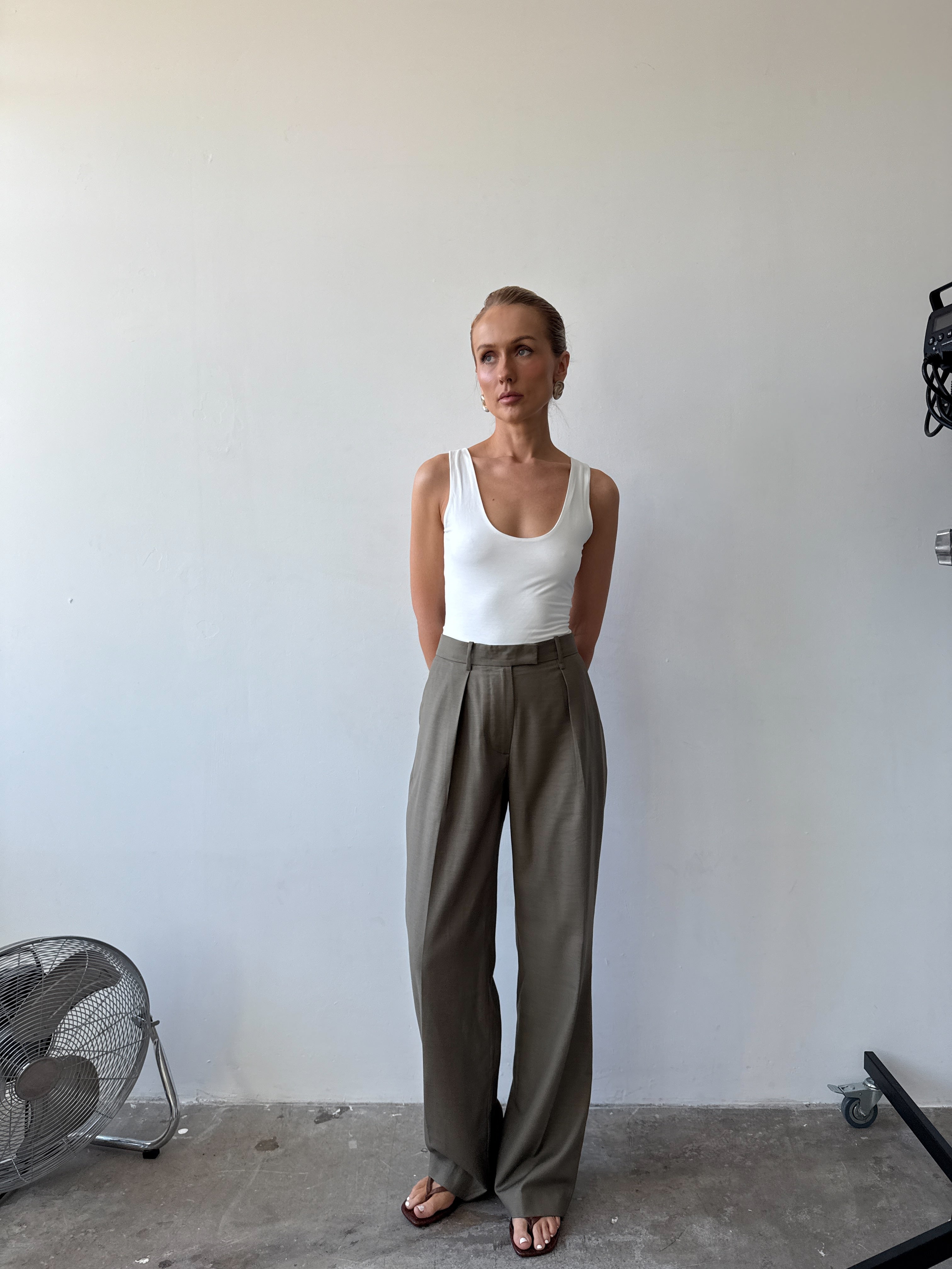 Model wearing a white tank top and light neutral green wide-leg pants with sandals standing against a plain wall.