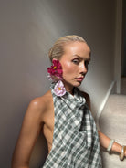 Woman with real flower earrings and a checkered draping top, sitting against a neutral background