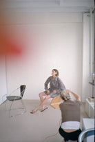 Two people in a room with minimal furniture, one photographs while the other poses wearing a plaid outfit
