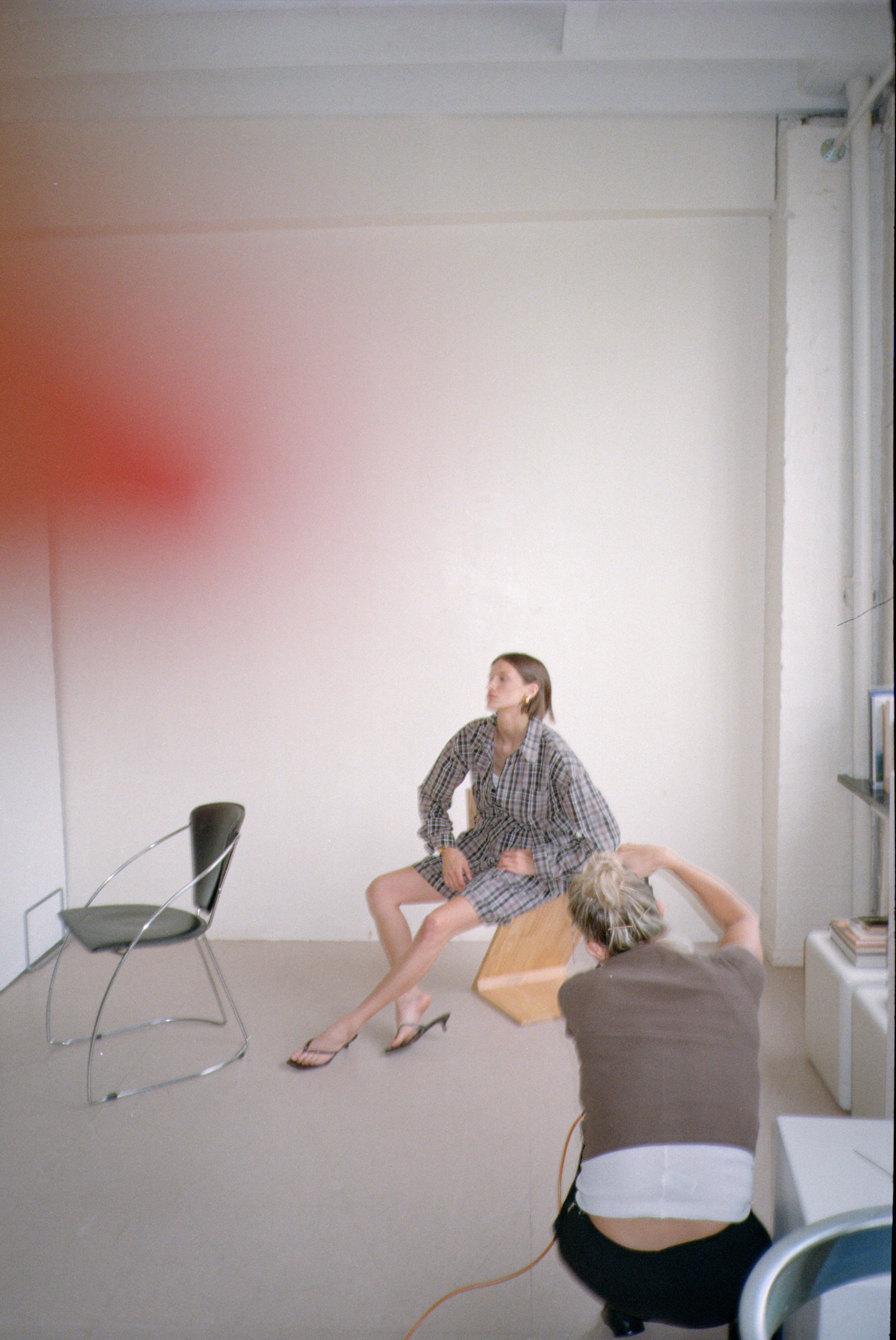 Two people in a room with minimal furniture, one photographs while the other poses wearing a plaid outfit