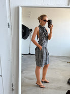 Model taking a mirror selfie wearing a plaid backless top with matching shorts accessorised by sunglasses and heeled sandals in a minimalistic room