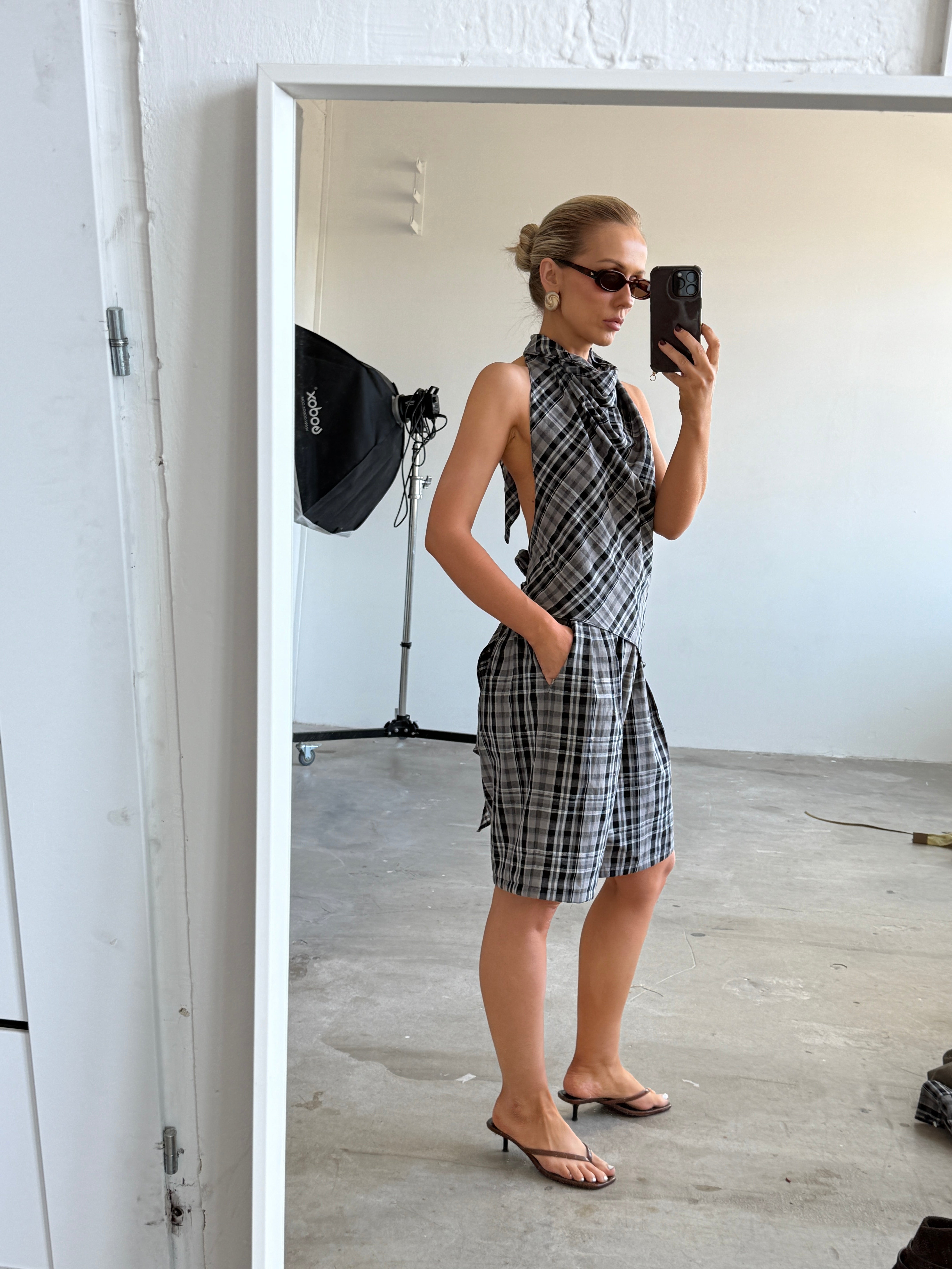 Model taking a mirror selfie wearing a plaid backless top with matching shorts accessorised by sunglasses and heeled sandals in a minimalistic room