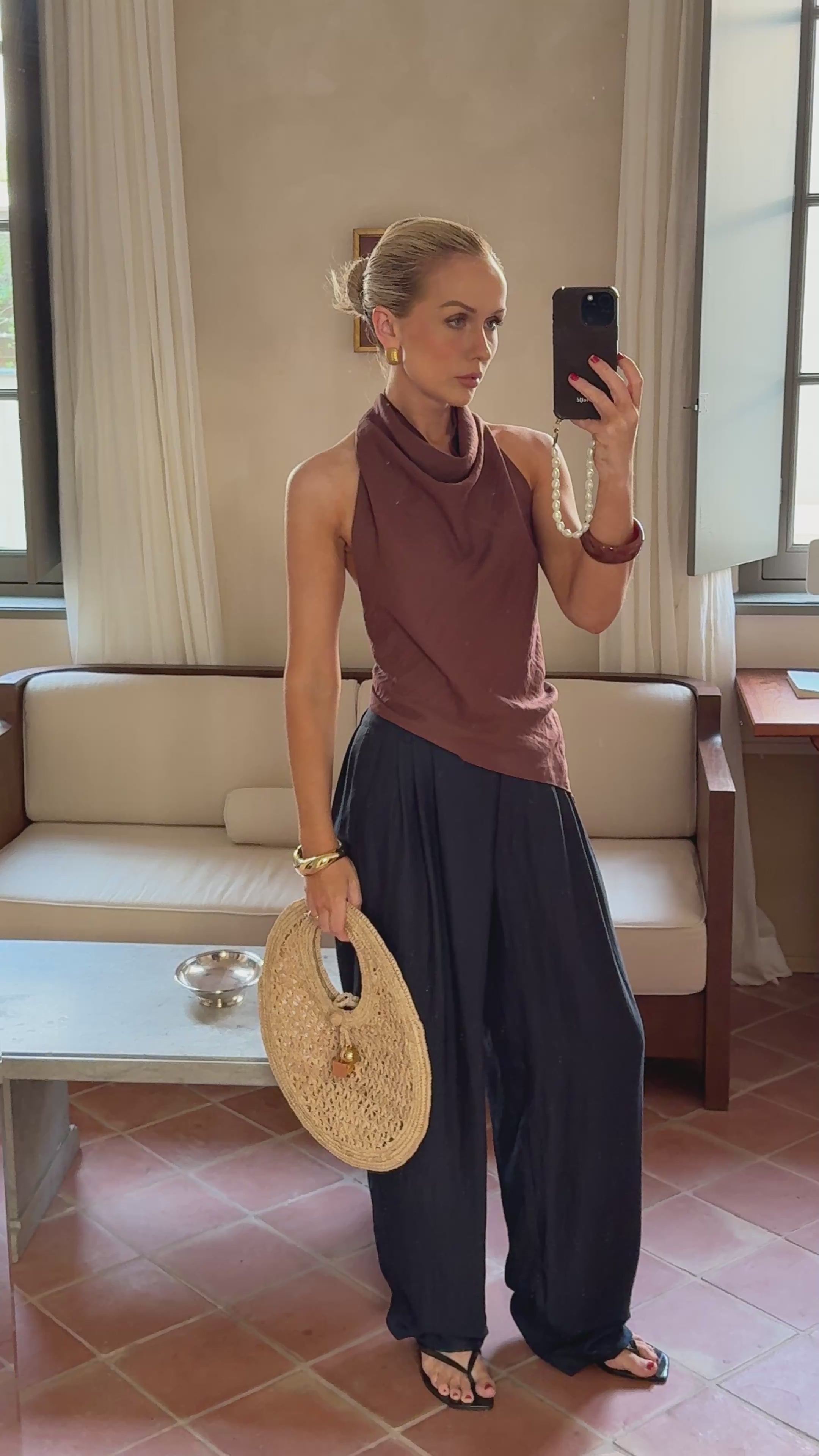 Model taking a mirror selfie while wearing a brown top and flowy navy trousers while holding a woven bag