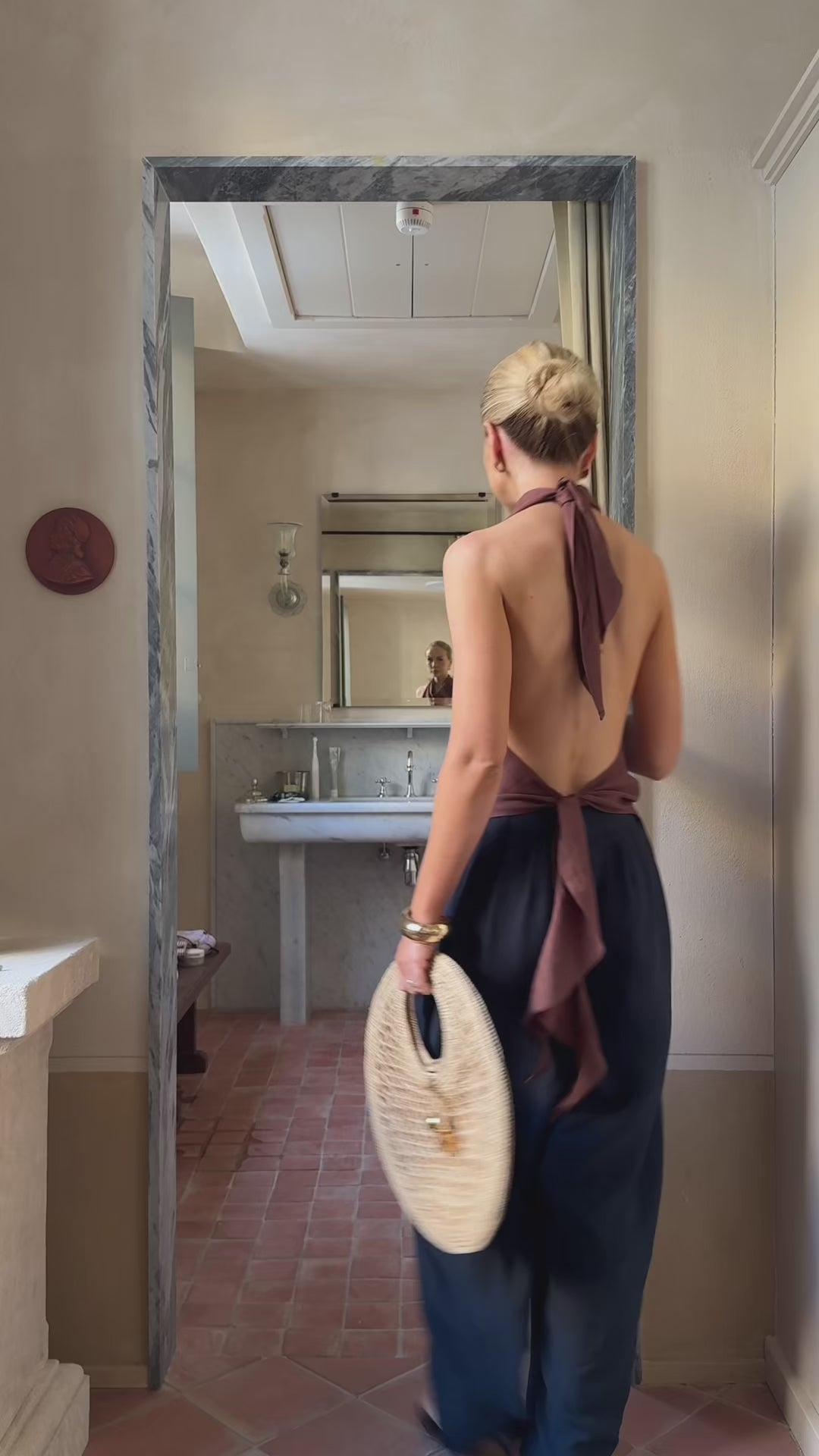 Model moves about a minimalistic room wearing an asymmetric halter neck top and black trousers while holding a straw bag
