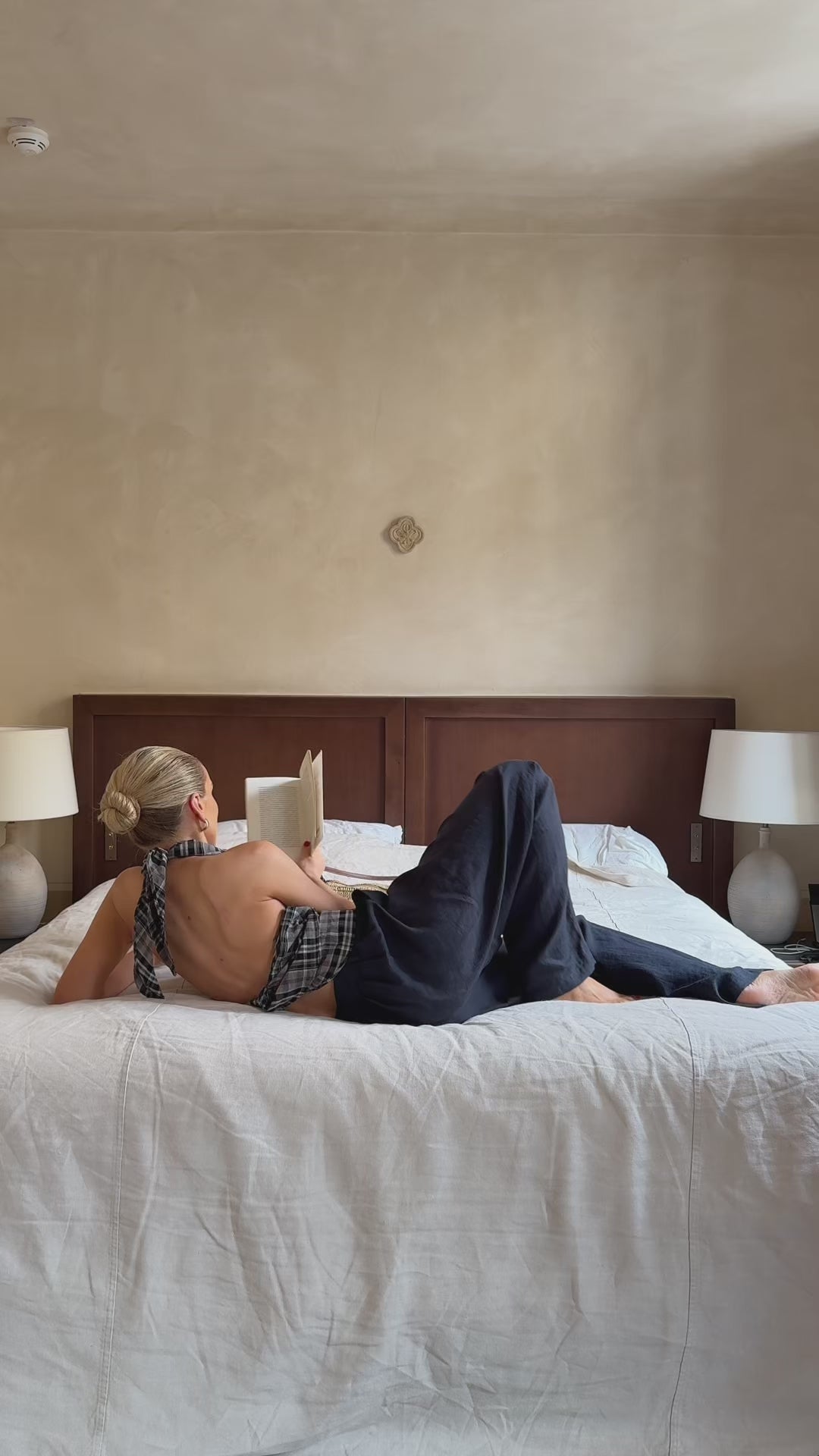 A woman reading on a bed, faced away from the camera. She's wearing an open back plaid top with navy trousers 