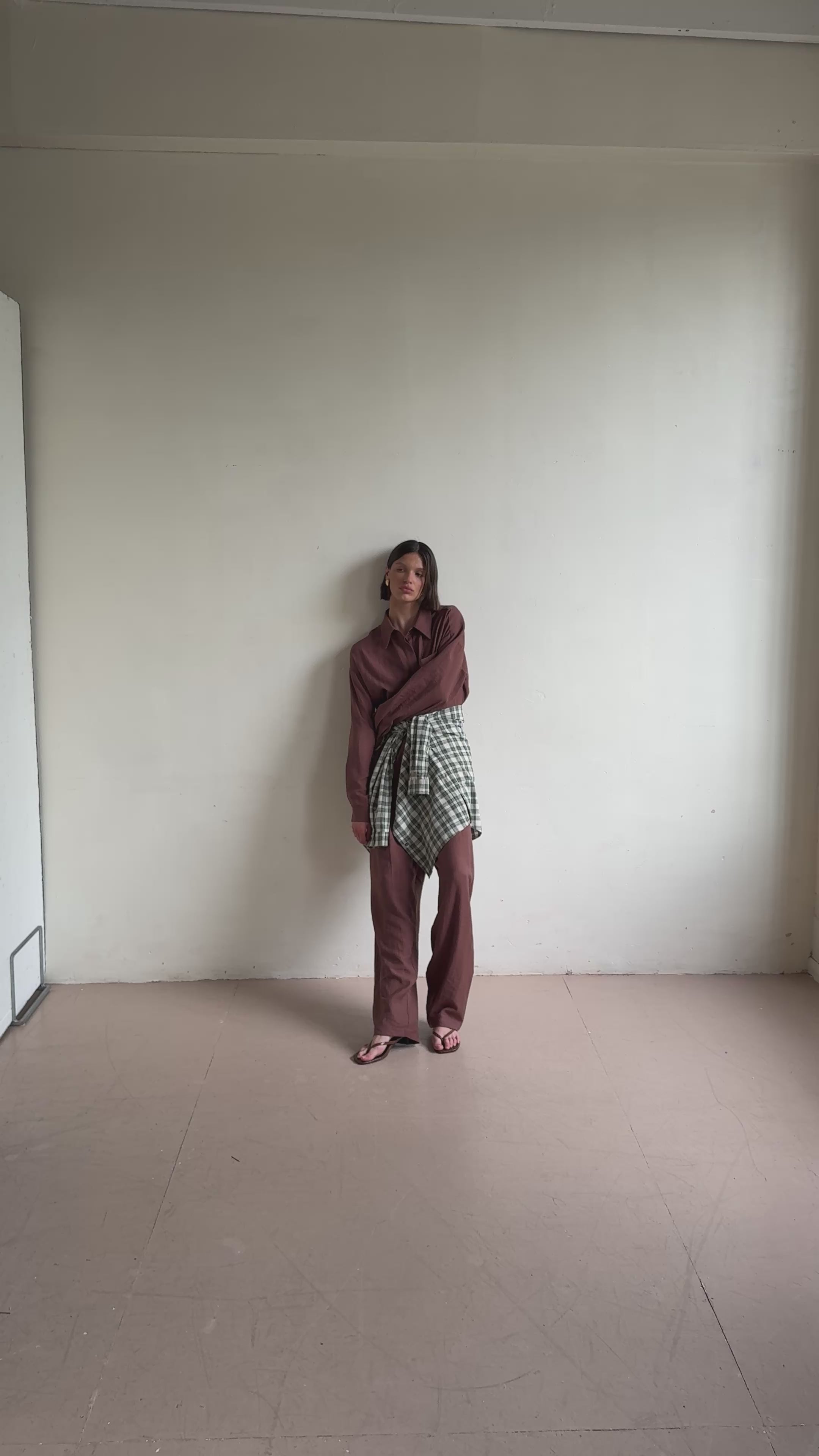Model posing against a white background while wearing a brown outfit and a white and green check shirt around the waist