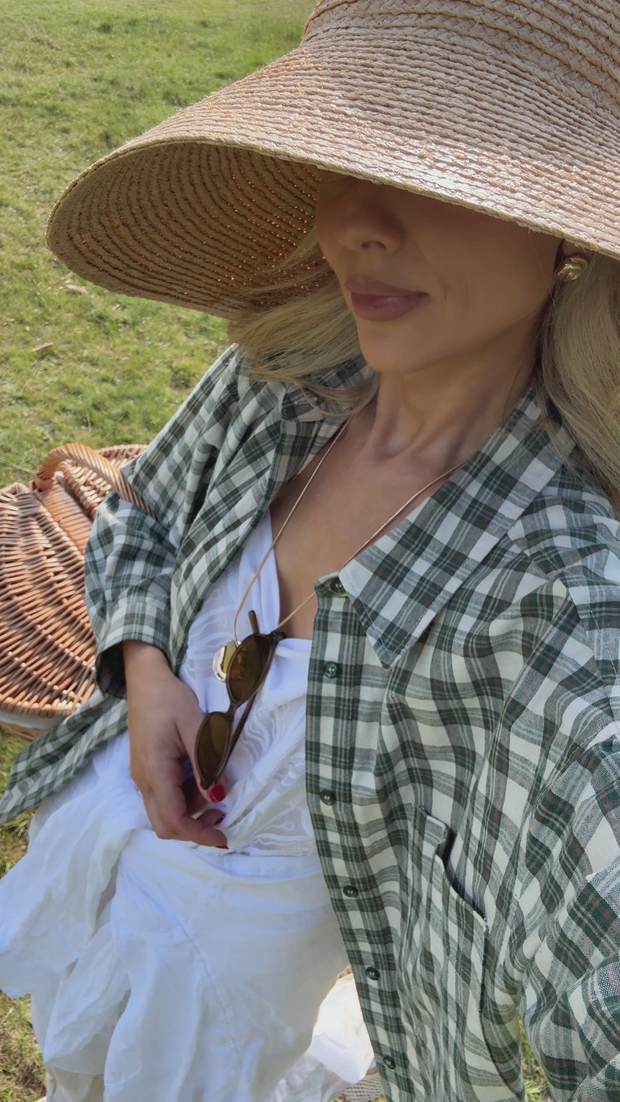 Woman walking in a field wearing a white outfit with a check shirt over the top and a wide brimmed woven hat