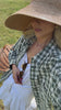 Woman walking in a field wearing a white outfit with a check shirt over the top and a wide brimmed woven hat