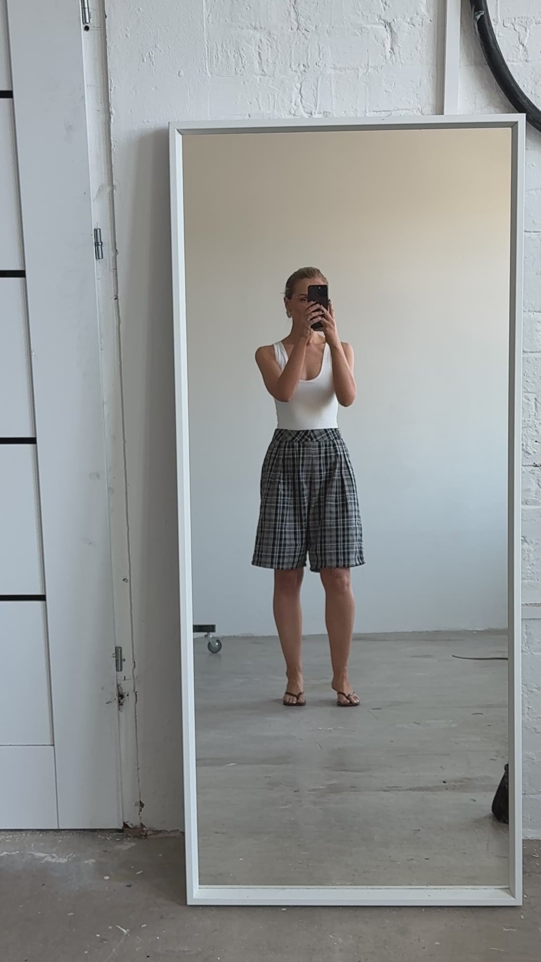 Model moves back and forth while taking a selfie, wearing a fitted white top and knee-length wide leg plaid shorts, demonstrating their comfort and ease of movement